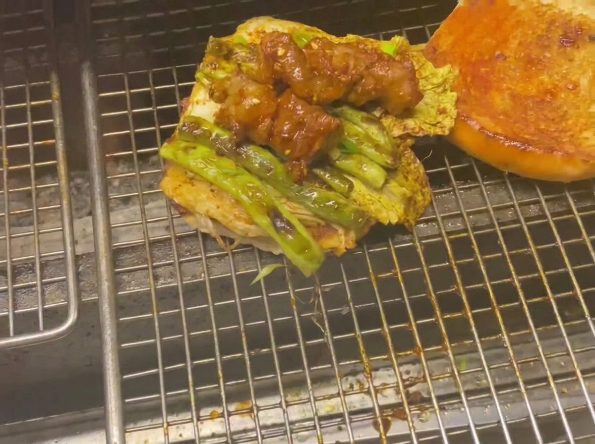 Bottom half of a burger bun topped with grilled vegetables and glazed meat skewers on a grill.