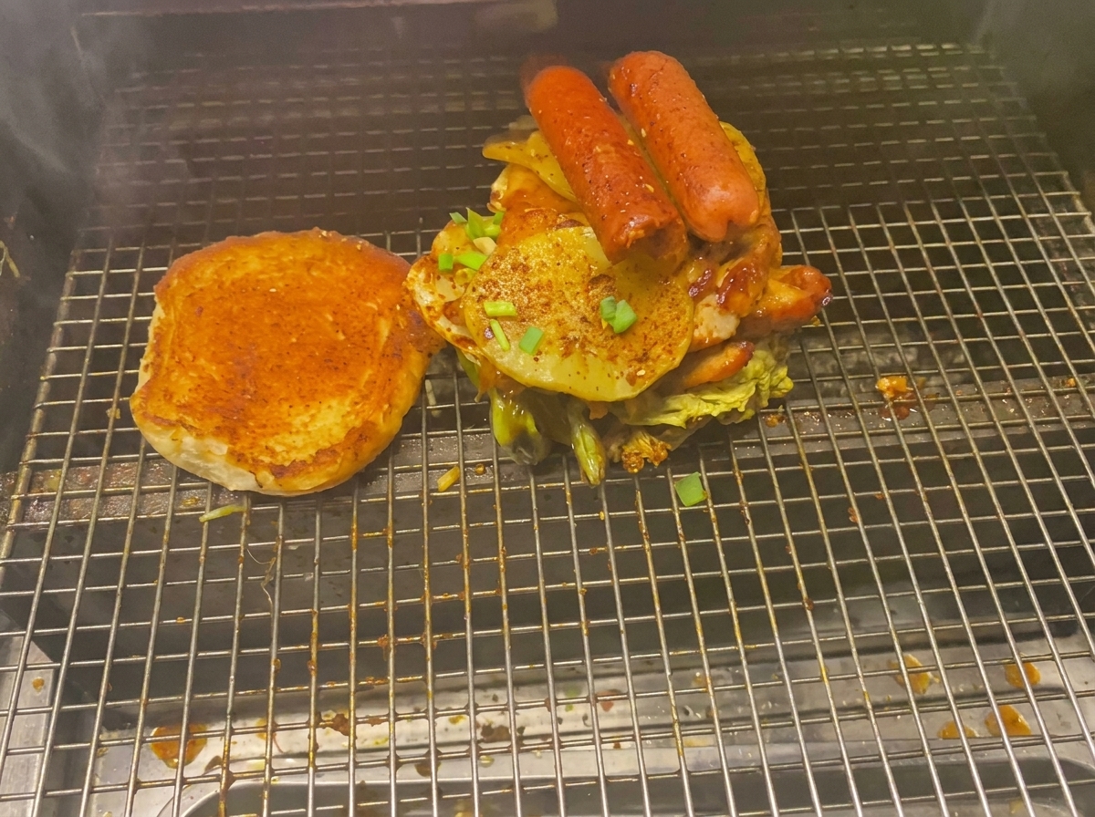 A towering burger loaded with meats, grilled potatoes, and sausages next to a toasted bun on a grill grate.