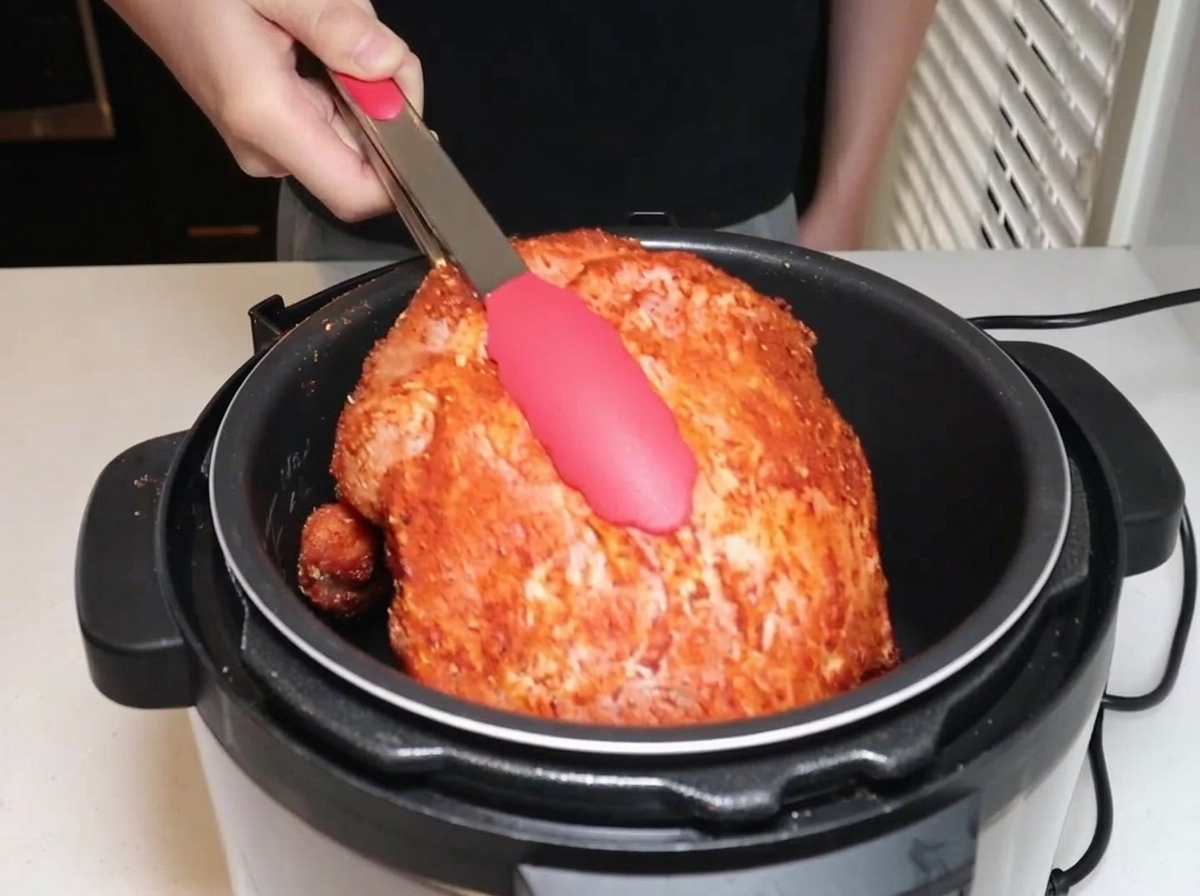 Seasoned pork shoulder being lowered into an electric pressure cooker with tongs.