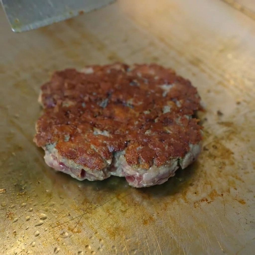 A slice of yellow cheese melting on a browned beef patty under a metal dome.