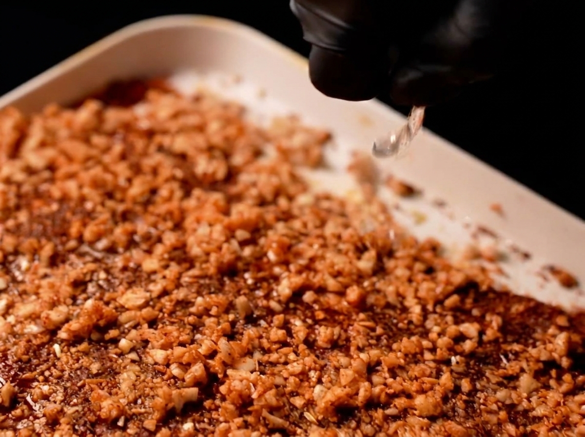 A splash of clear liquid being poured by gloved fingers over garlic-covered pork ribs.