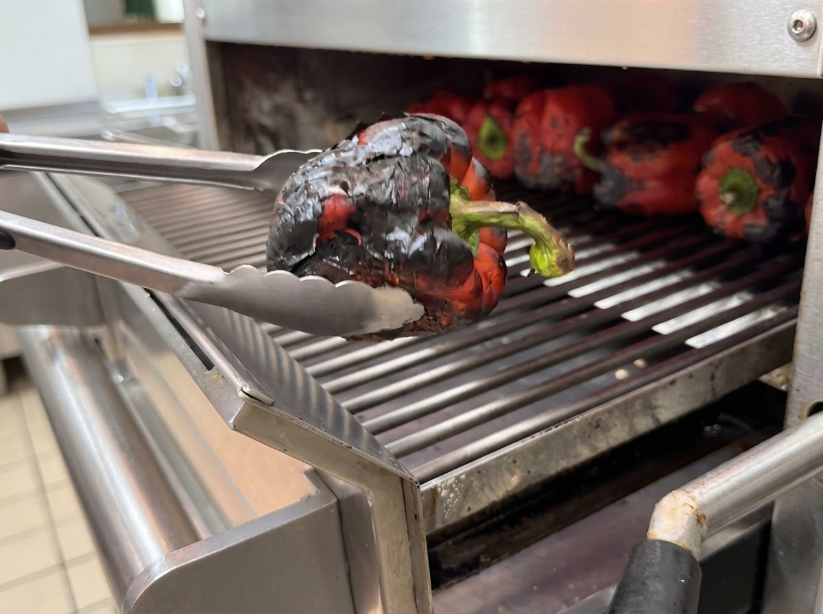 Charred bell peppers inside a container being covered with plastic wrap to steam.