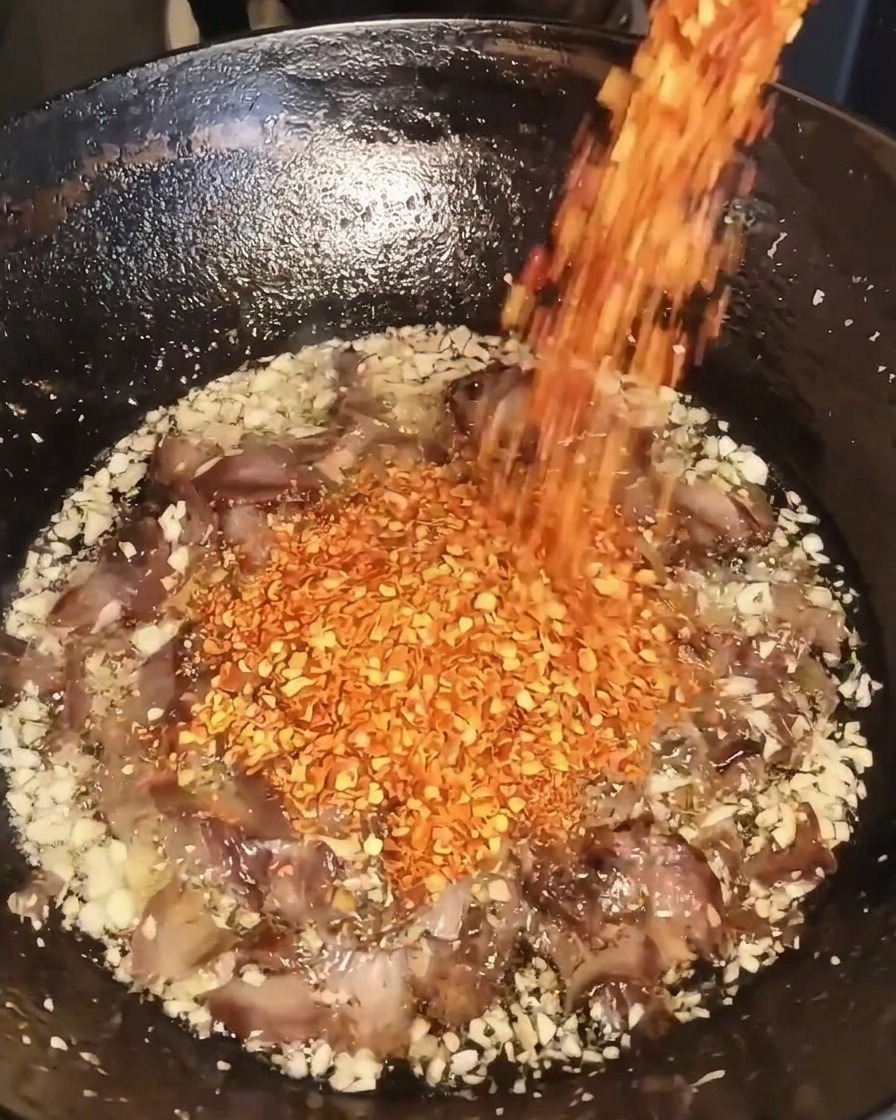 Dry red chili flakes being poured from a container into a wok containing fried garlic, beef, and oil.