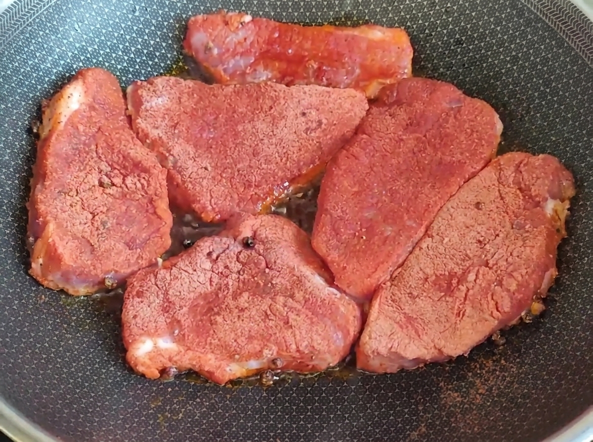 Thick slices of raw pork coated in red seasoning searing in a hot pan.