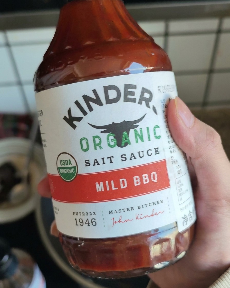 A hand holding a bottle of mild BBQ sauce, preparing to add it to the shredded pork.