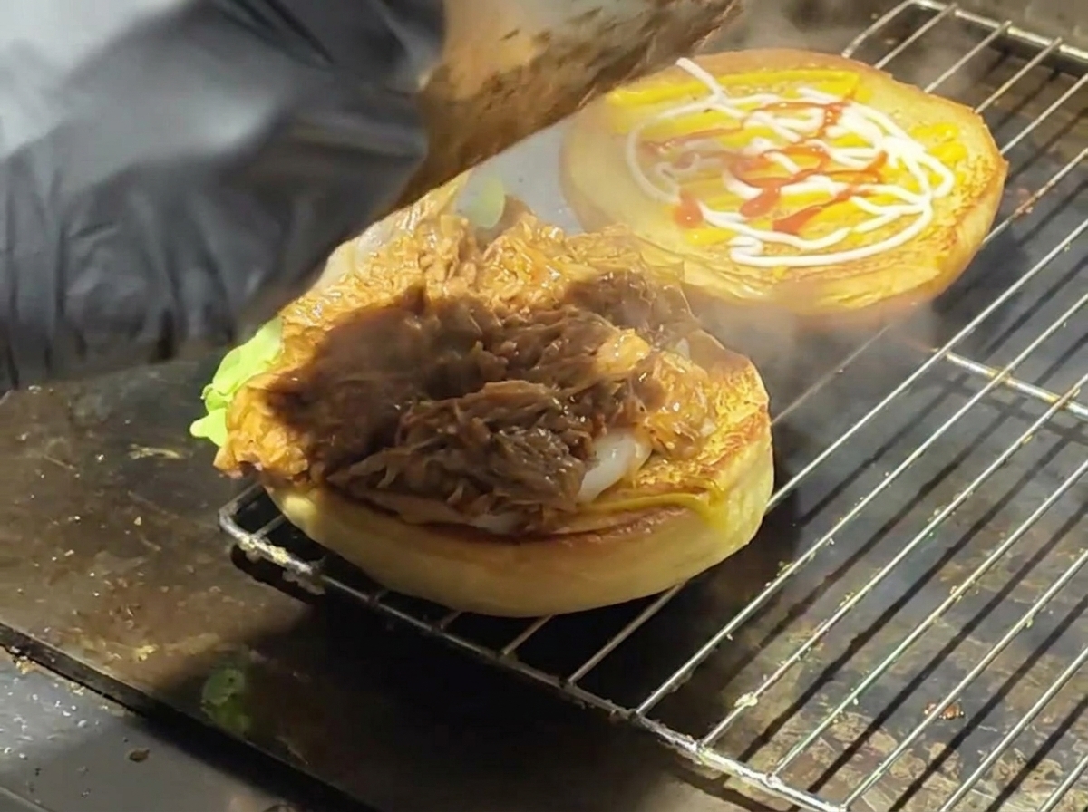 Saucy pulled pork being scooped onto a toasted bun with lettuce on a flat top grill.