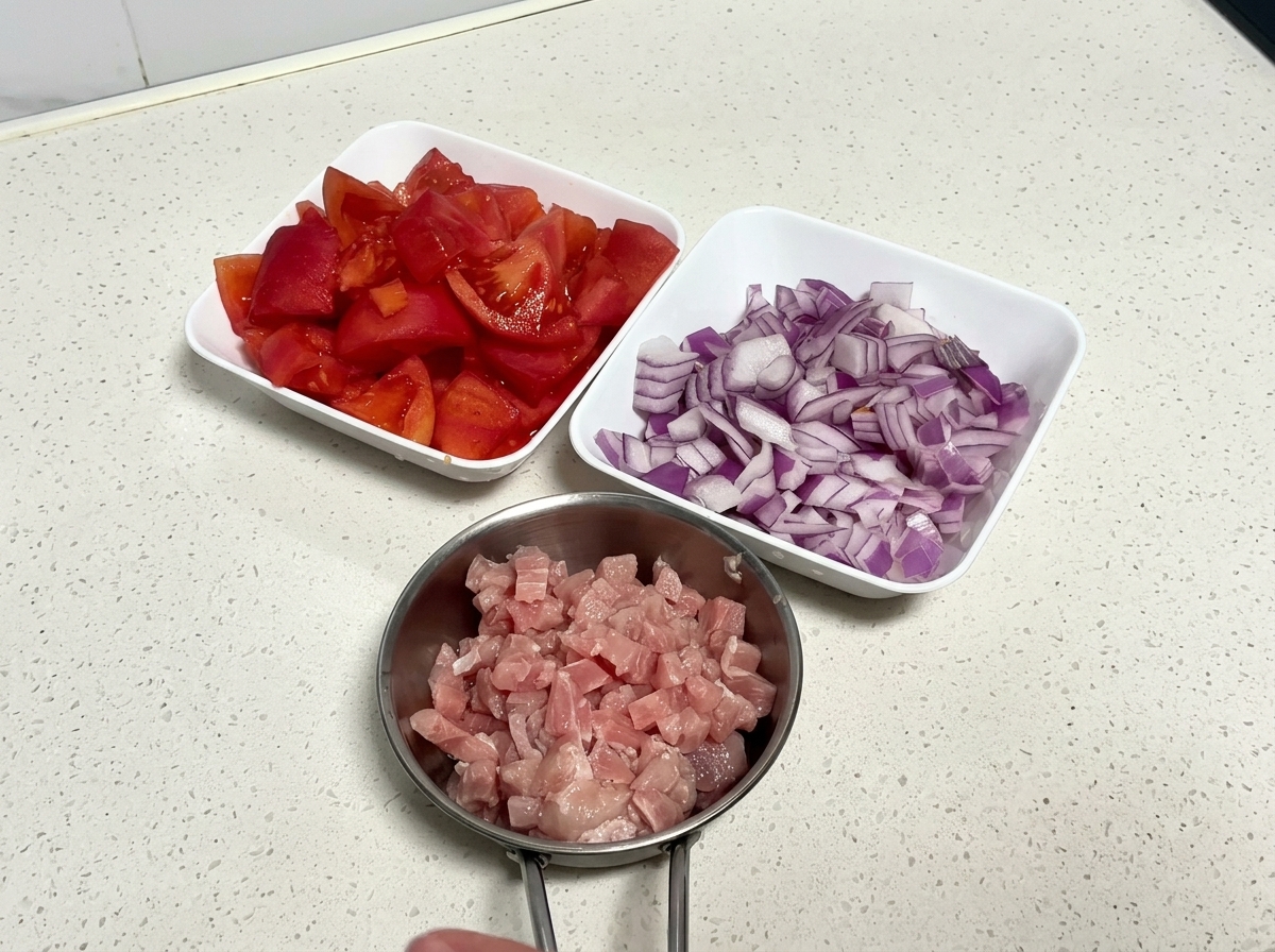 A top-down view of three white bowls filled with raw diced tomatoes, chopped red onion, and raw cubed pork on a clean countertop.