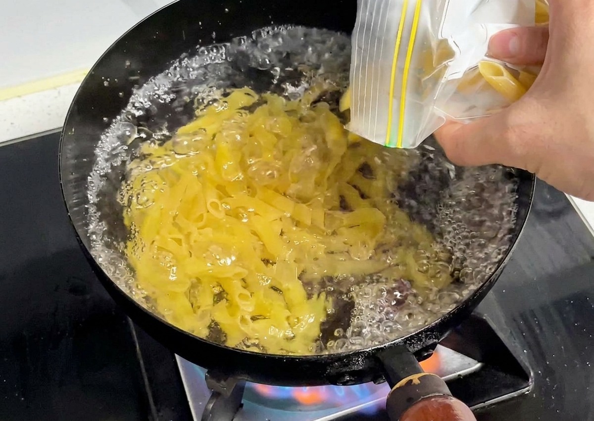 Dry yellow penne pasta being poured into a pot of vigorously boiling water.