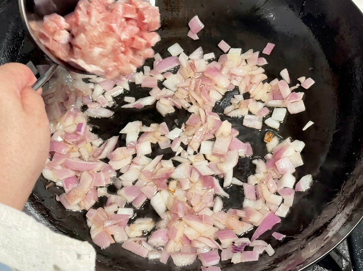 Raw cubed pork being tipped from a metal bowl into a wok filled with sautéed red onions.