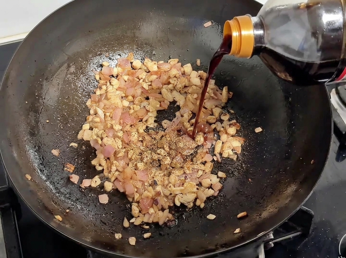 Soy sauce being poured from a bottle into a wok containing browned pork and onions, topped with black pepper.