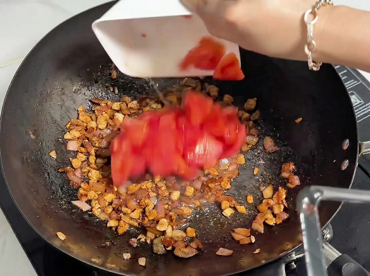 Freshly chopped red tomatoes being added from a white bowl into a wok containing cooked minced meat.