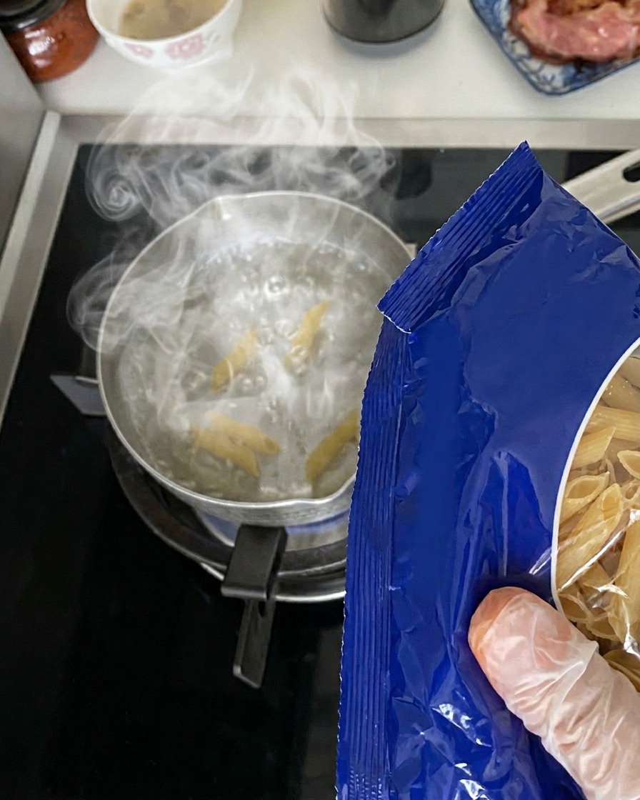 Dry penne pasta being poured from a blue package into a metal pot of boiling water on a stovetop.