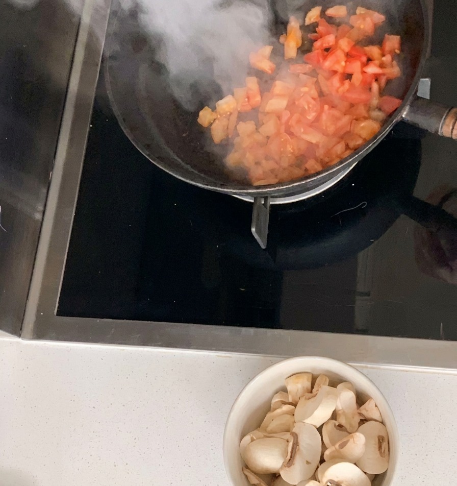 Diced tomatoes being stir-fried in a black skillet with a bowl of sliced white mushrooms resting on the counter below.