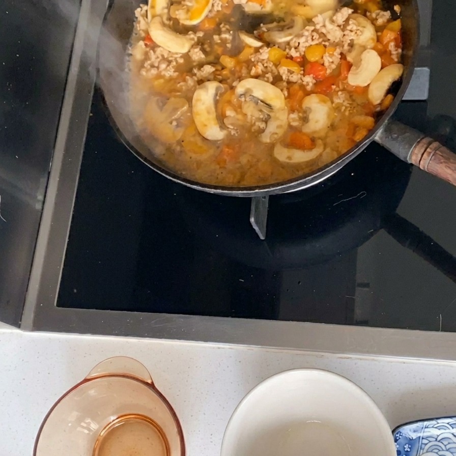 A simmering sauce made of minced pork, sliced mushrooms, and diced tomatoes bubbling in a black frying pan.