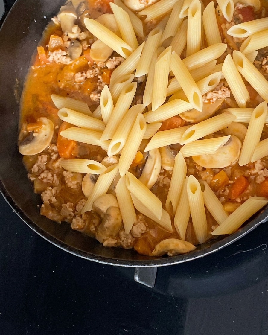 Cooked penne pasta resting on top of a savory minced meat, mushroom, and tomato sauce in a black skillet.