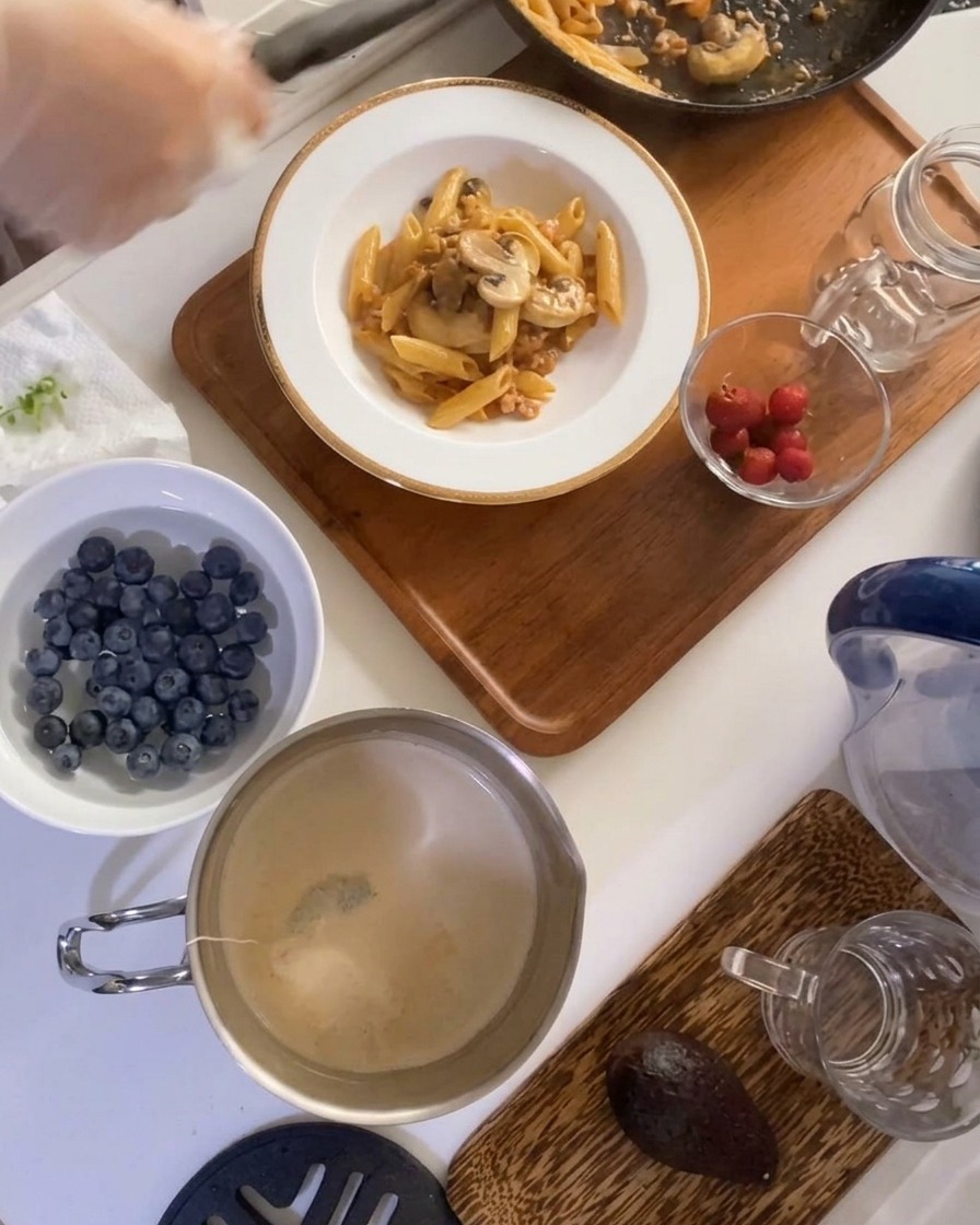 A portion of macaroni with meat and mushroom sauce plated in a deep white bowl on a wooden tray, with fresh fruits nearby.