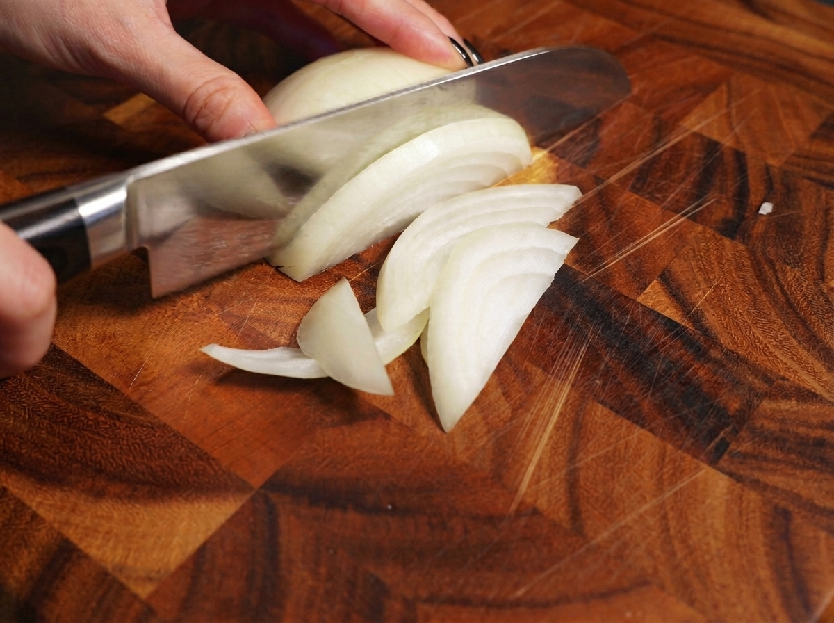 A hand using a chefs knife to slice a white onion on a wooden cutting board.