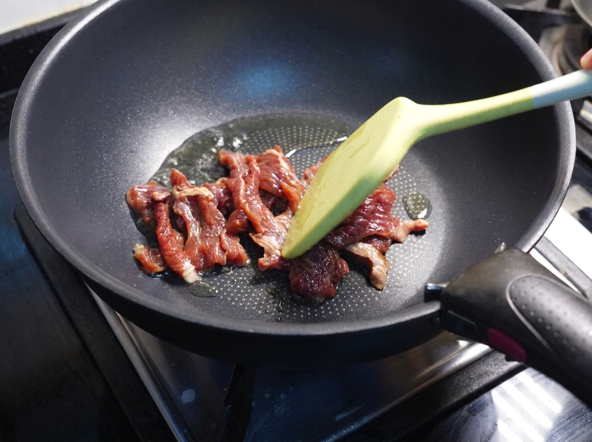 Spatula stir-frying partially cooked beef slices in a dark pan.