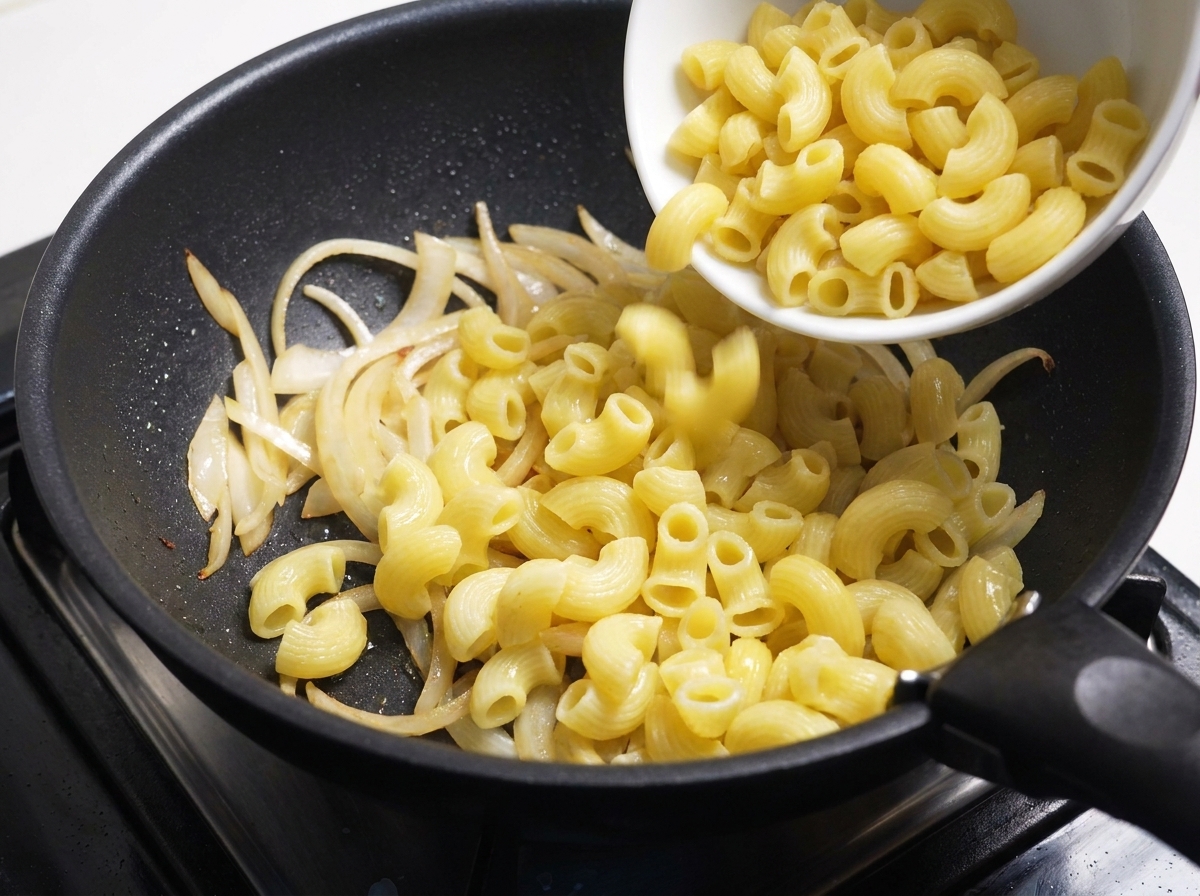 Cooked macaroni being poured from a white bowl into a pan of sautéed onions.