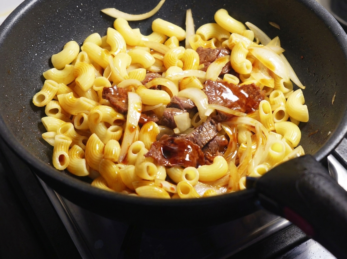 Dark soy sauce poured over macaroni, beef slices, and onions in a black frying pan.