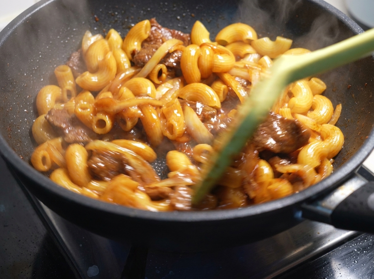 Macaroni, beef, and onions being rapidly tossed and stirred with a spatula in a hot pan.