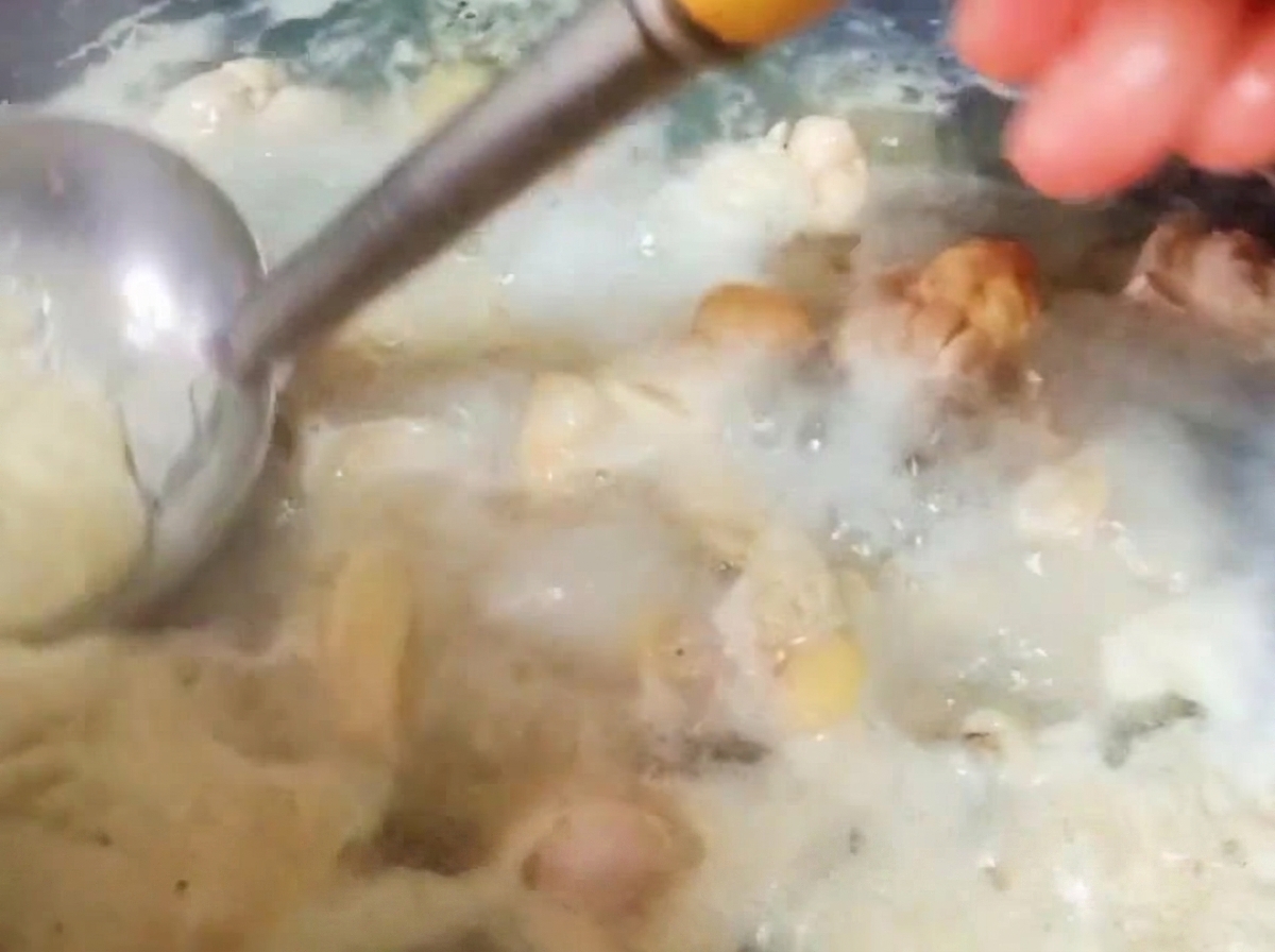 A metal ladle skimming thick blood foam off the surface of boiling water containing chicken pieces.