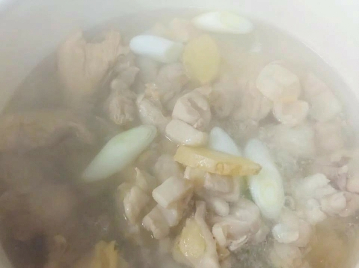 Hot water actively steaming in a white pot over chicken pieces, sliced ginger, and scallions.