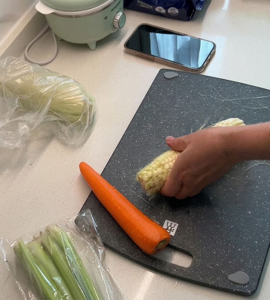 Fresh corn kernels being sliced off the cob on a cutting board, with the remaining husk and silk set aside.
