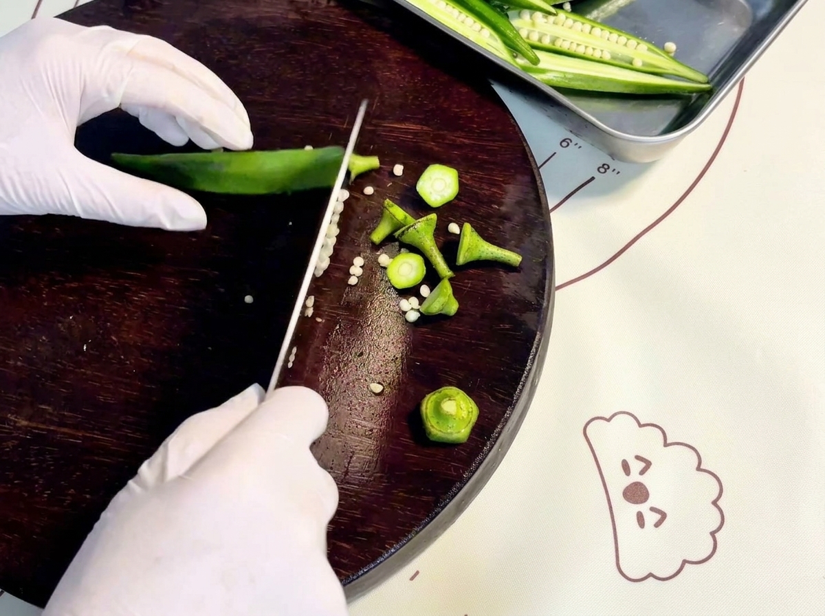 Gloved hands slicing fresh green okra lengthwise on a dark wooden cutting board.