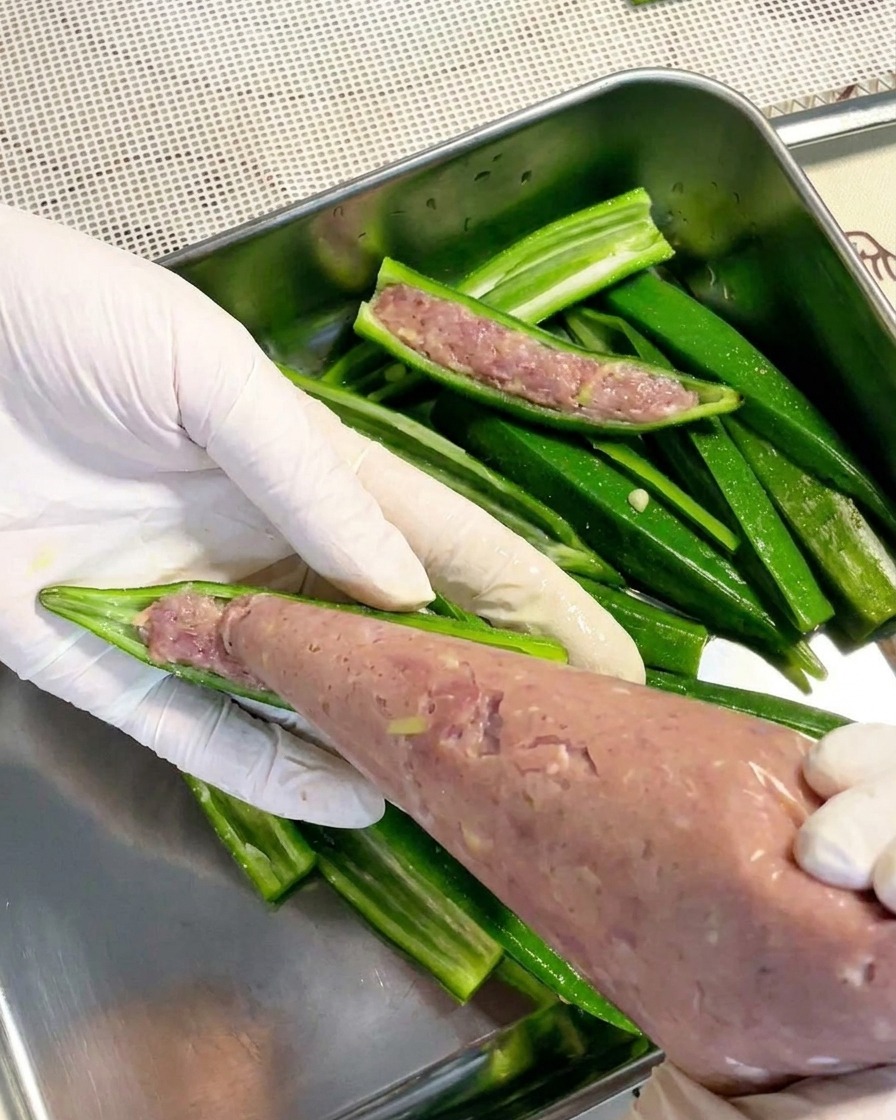 A pair of gloved hands using a clear piping bag to fill a blanched okra half with a smooth, light-colored meat paste.