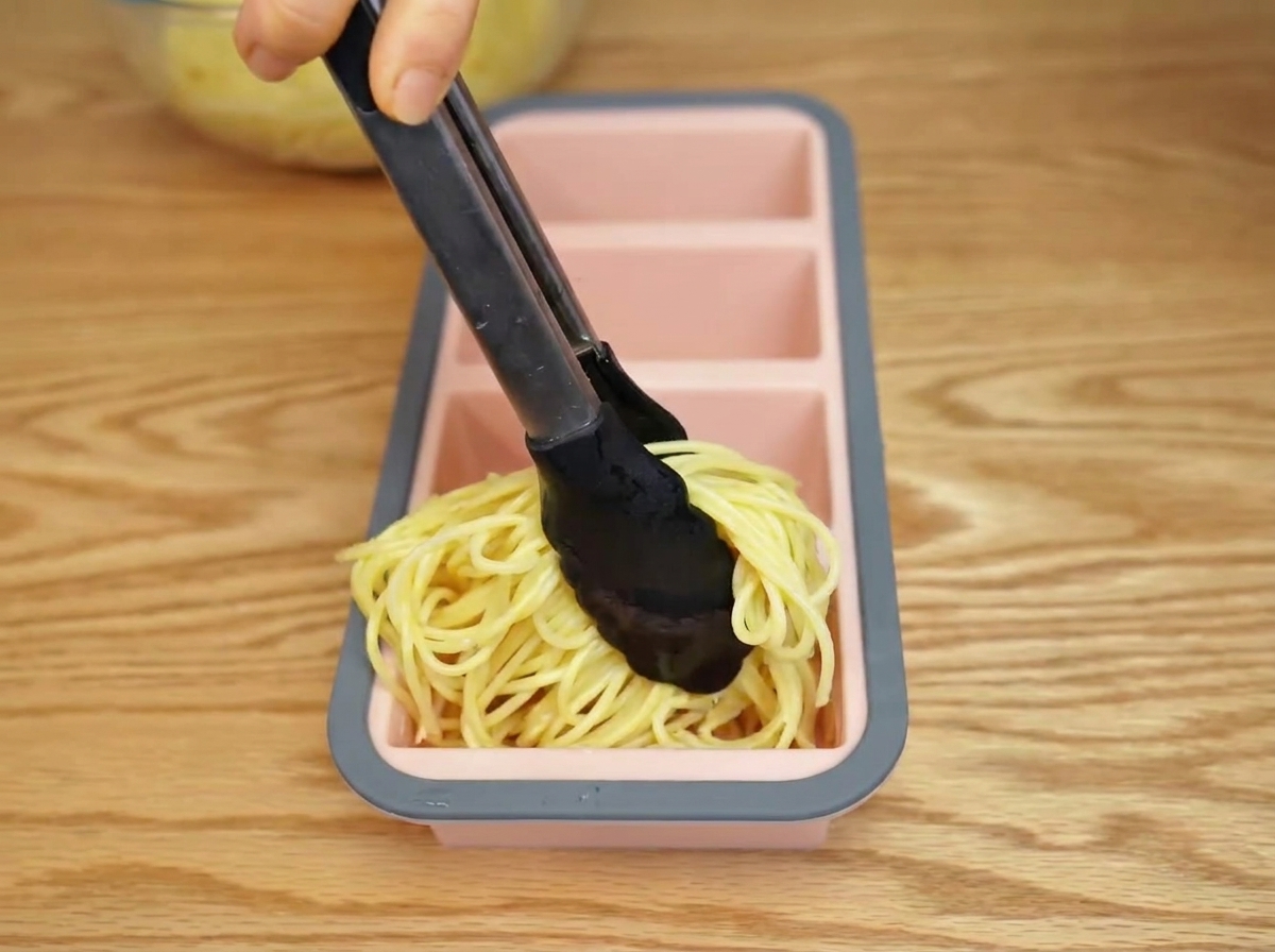Cooked spaghetti being placed into a silicone mold with tongs.