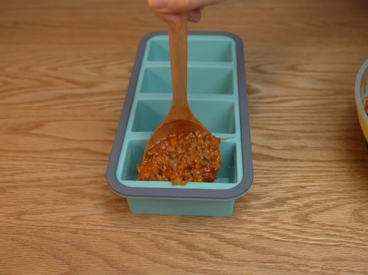 Thick tomato and ground beef sauce being spooned into individual compartments of a blue silicone tray.