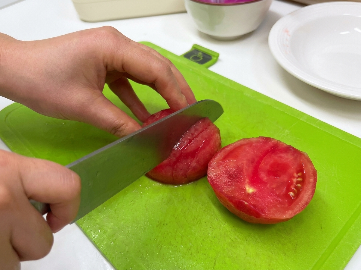 A person using a large kitchen knife to slice a peeled red tomato on a green cutting board.