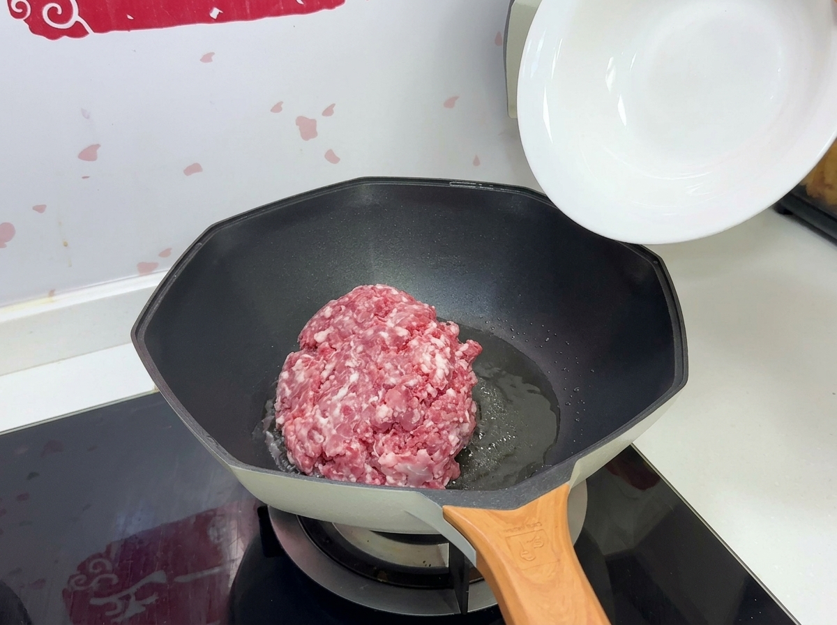 A mound of raw minced pork being dropped from a white bowl into a hot pan with oil.