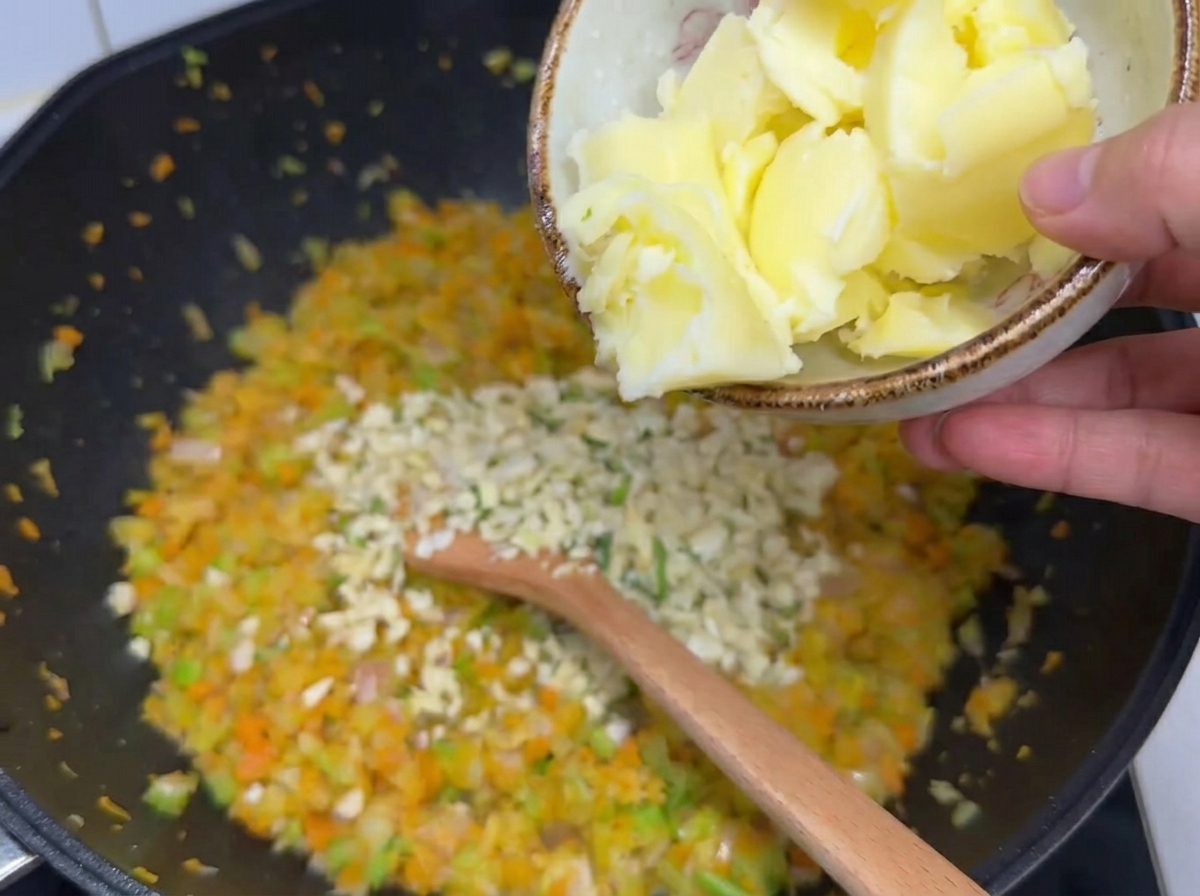 Large chunks of butter being poured from a small bowl over softened chopped vegetables and minced garlic in a wok.