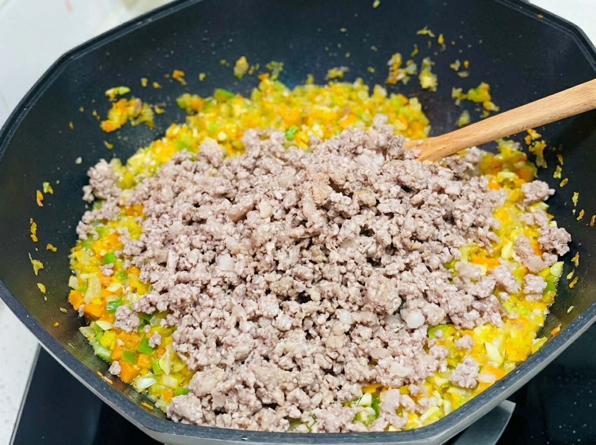 Minced pork being stirred into a mixture of sautéed diced carrots, celery, and onions in a large wok.