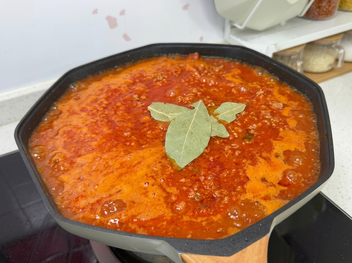 A simmering tomato meat sauce in a wok, seasoned with salt, sugar, bay leaves, and black pepper.