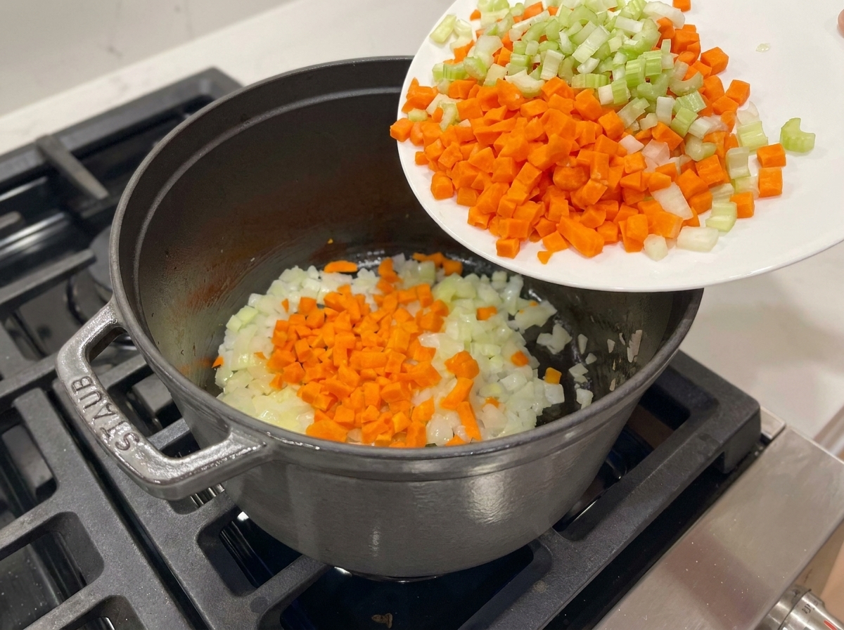 Diced carrots and celery being poured from a white plate into a pot with sautéed onions.