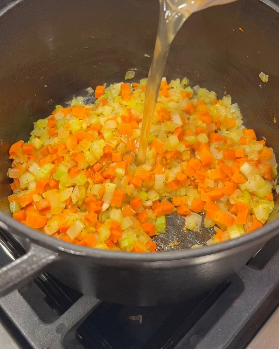 Golden chicken broth being poured from a glass measuring cup into a pot containing sautéed onions, carrots, and celery.