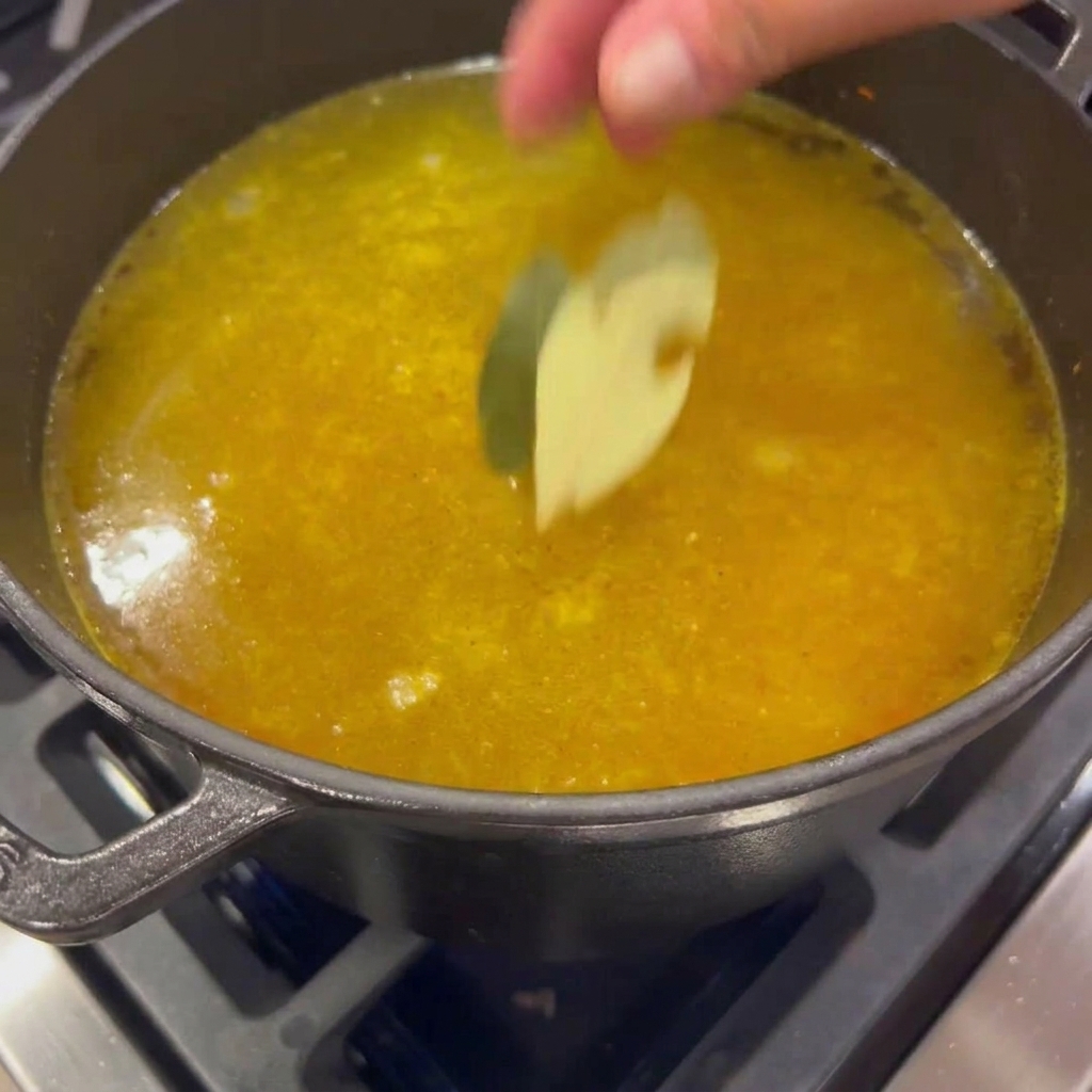 Two dried bay leaves being dropped into a pot of simmering, golden vegetable broth.