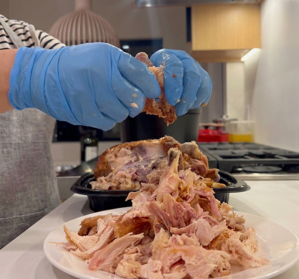 Hands wearing blue culinary gloves shredding cooked rotisserie chicken meat into a pile on a white plate.