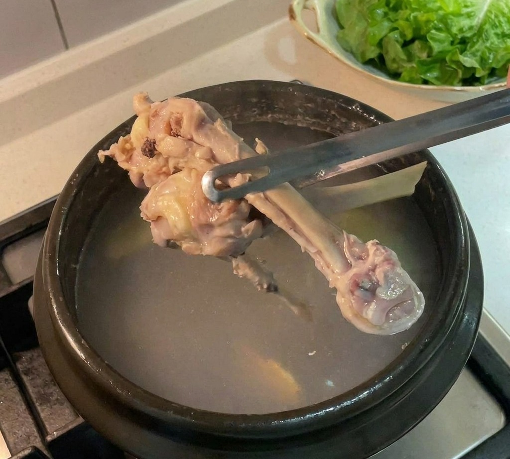 Chicken bones being lifted out of a dark pot of broth using metal tongs.