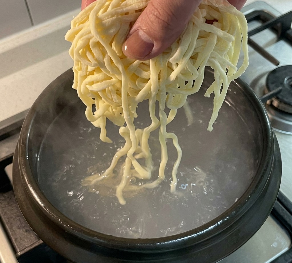 Fresh noodles being lowered into a pot of boiling chicken broth.