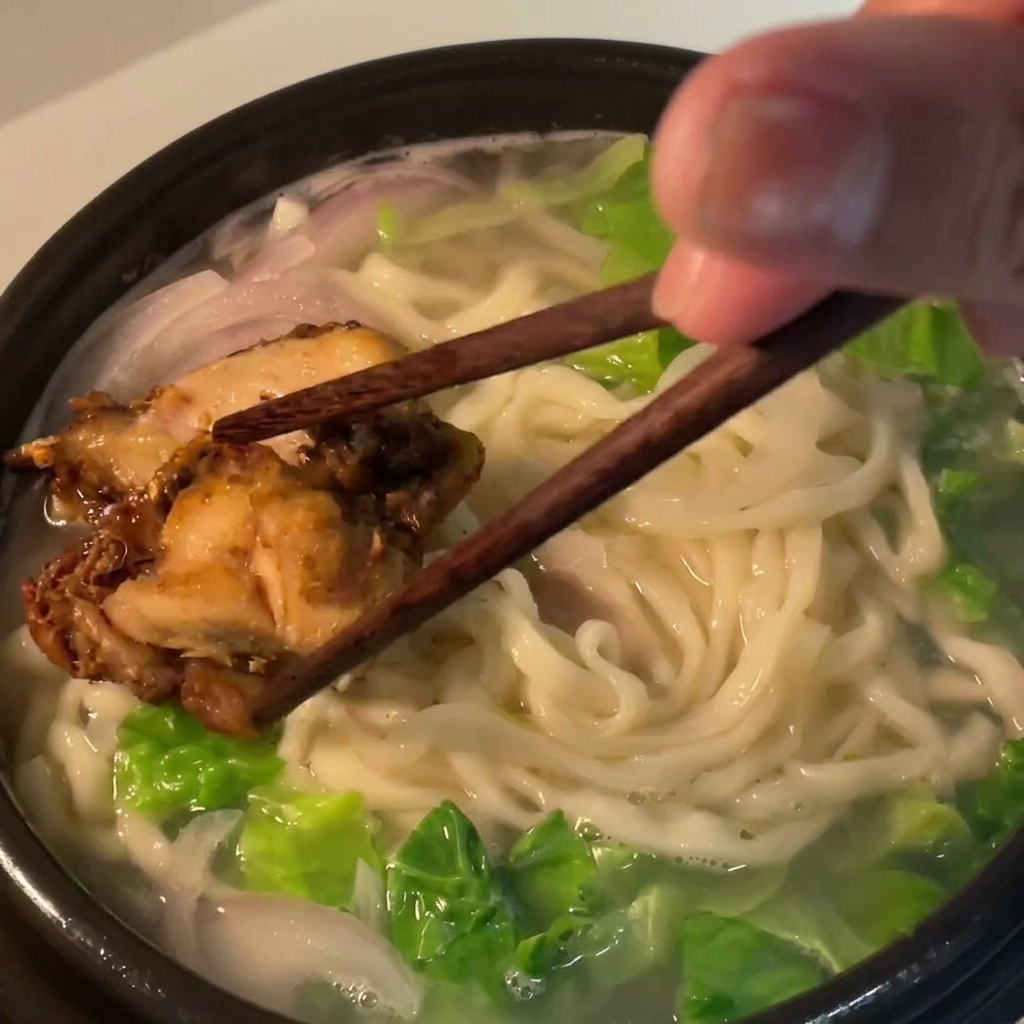 Cooked chicken thigh pieces being placed onto the bed of noodles in the soup bowl using dark wooden chopsticks.