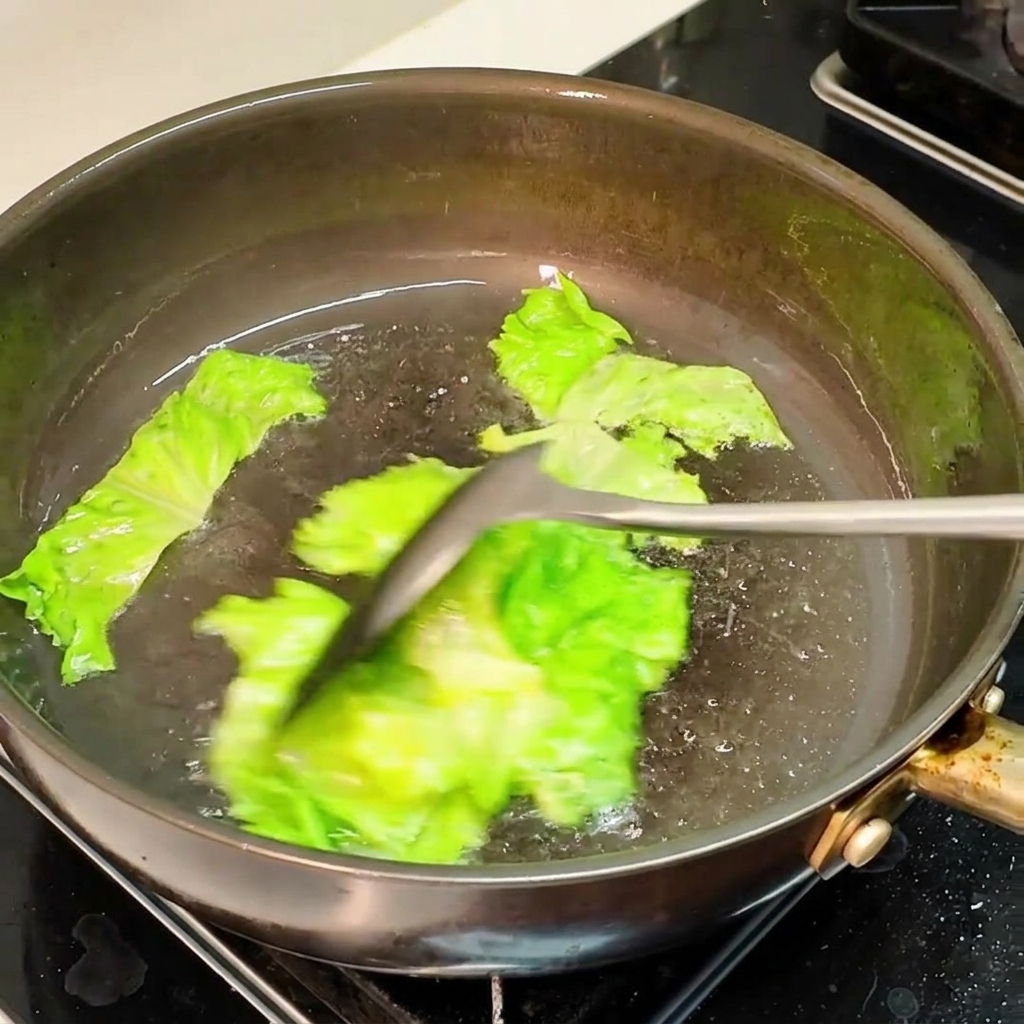 Pieces of green lettuce being actively stir-fried in a hot oiled pan with a spatula.