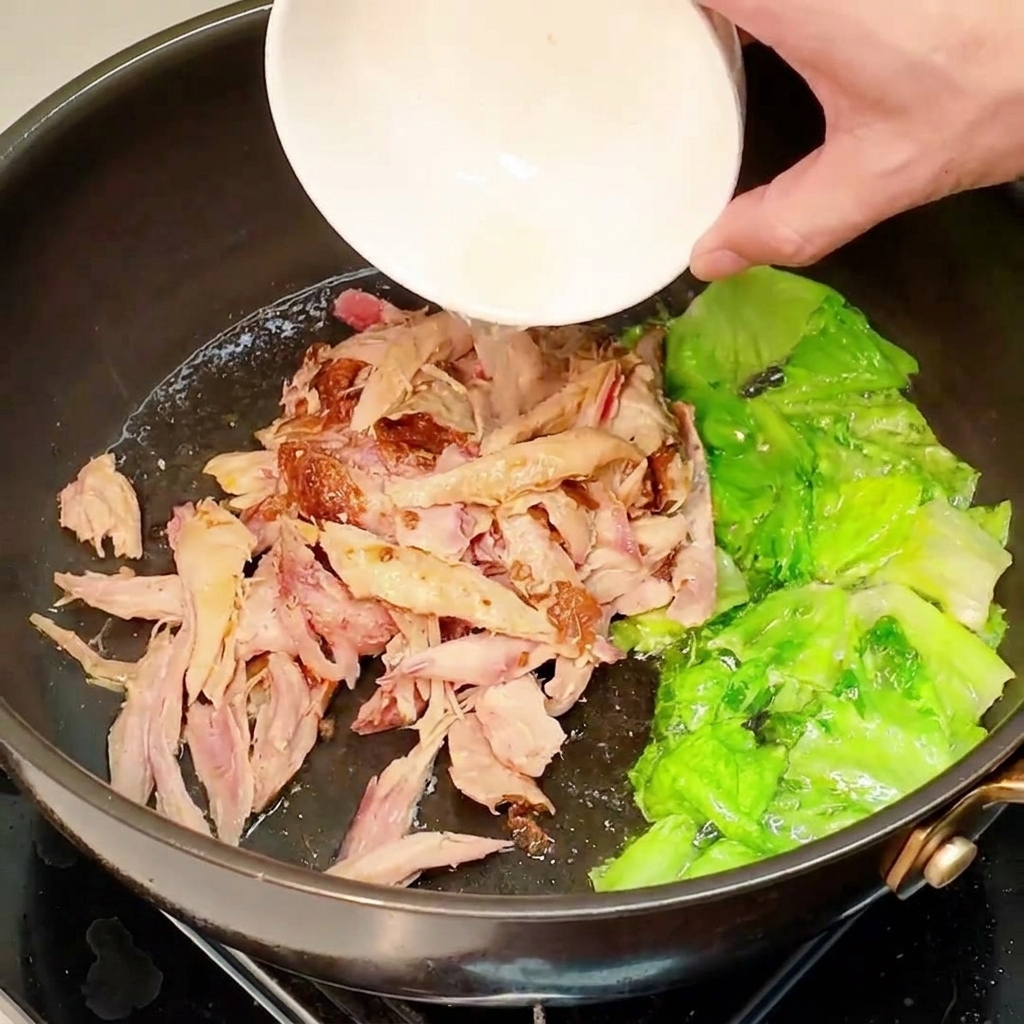 Shredded pre-cooked chicken being poured from a small white bowl into a pan containing stir-fried lettuce.