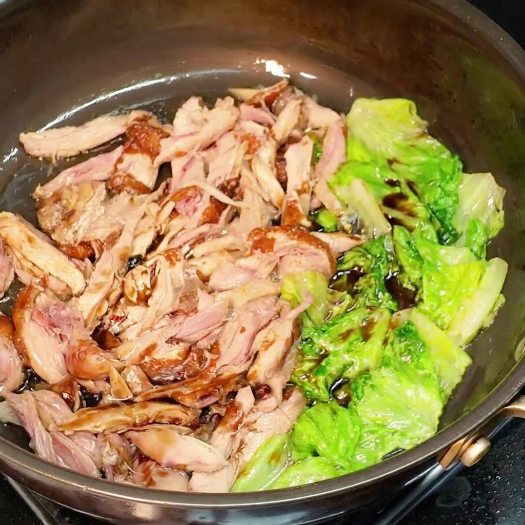 Shredded chicken and lettuce coated in dark soy sauce being mixed together in a pan.