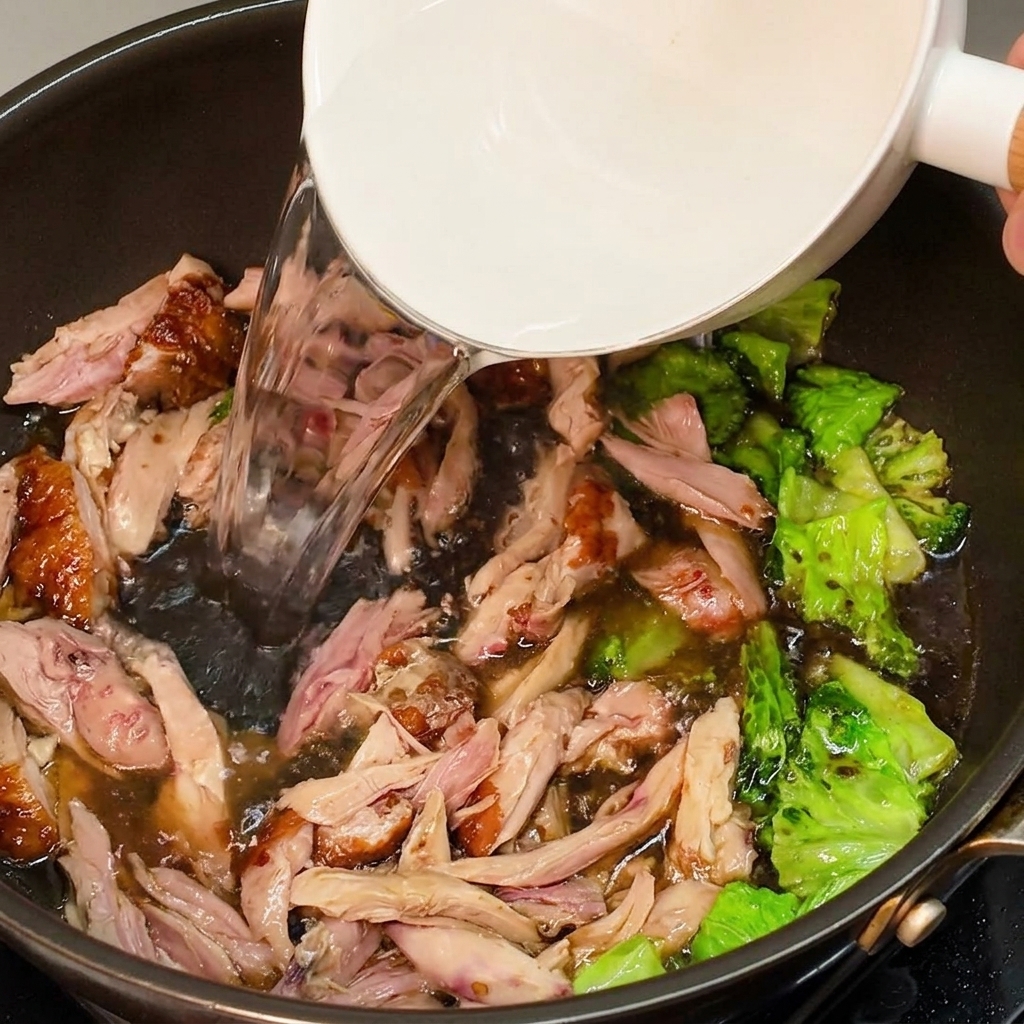 Clear hot water being poured from a white bowl into a pan with seasoned chicken and greens to form a broth.