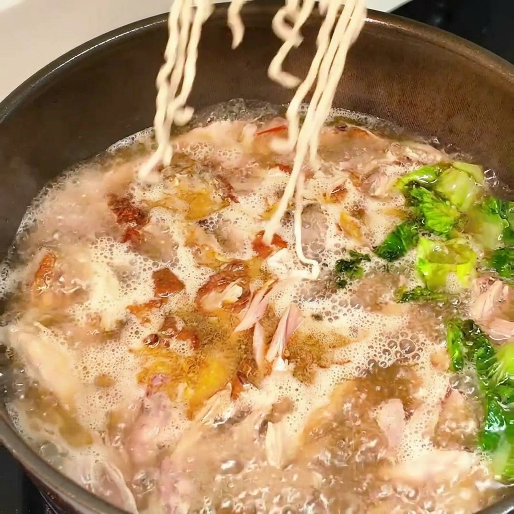 A block of wavy dry noodles being lowered into a pan of bubbling hot chicken and lettuce broth.