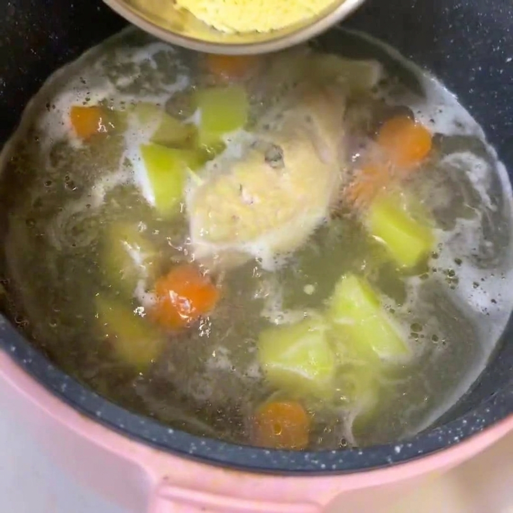 A pot of simmering broth containing chicken, diced potatoes, and carrots, with baby noodles being stirred in.